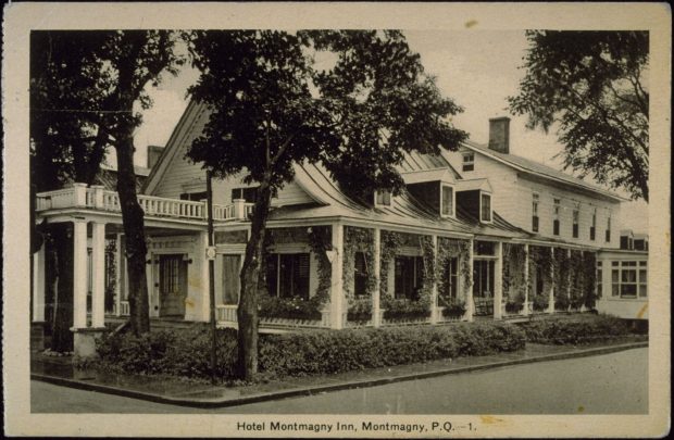 Photographie d’une carte postale en noir et blanc où l’on voit l’hôtel Montmagny Inn situé au coin de deux rues. Il s’agit d’un bâtiment d’un étage et demi avec une toiture courbée percée de trois lucarnes. L’immeuble est ceinturé d’une galerie munie de son propre toit. L’annexe, juxtaposée à droite, possède deux étages avec un toit à deux versants droits.