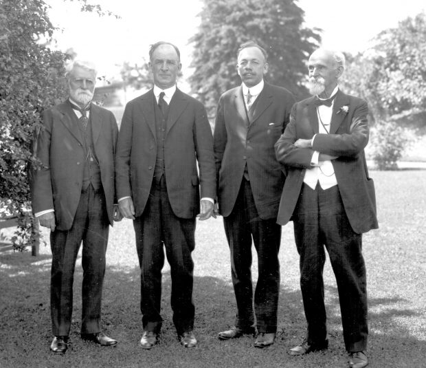 Photographie en noir et blanc de quatre messieurs bien habillés vers 1920, debout sur une pelouse.