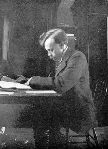 Photographie en noir et blanc d’un homme assis à un bureau sur une chaise en bois et regardant des documents sur son bureau.