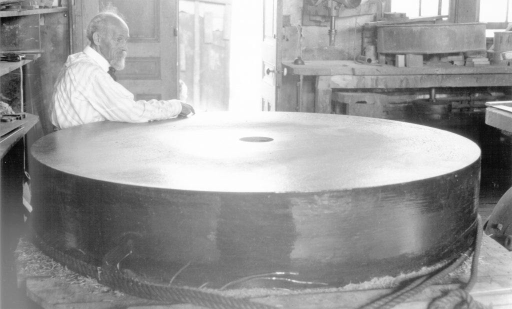 Photographie en noir et blanc d’un homme s’appuyant sur un grand morceau de verre épais et circulaire.