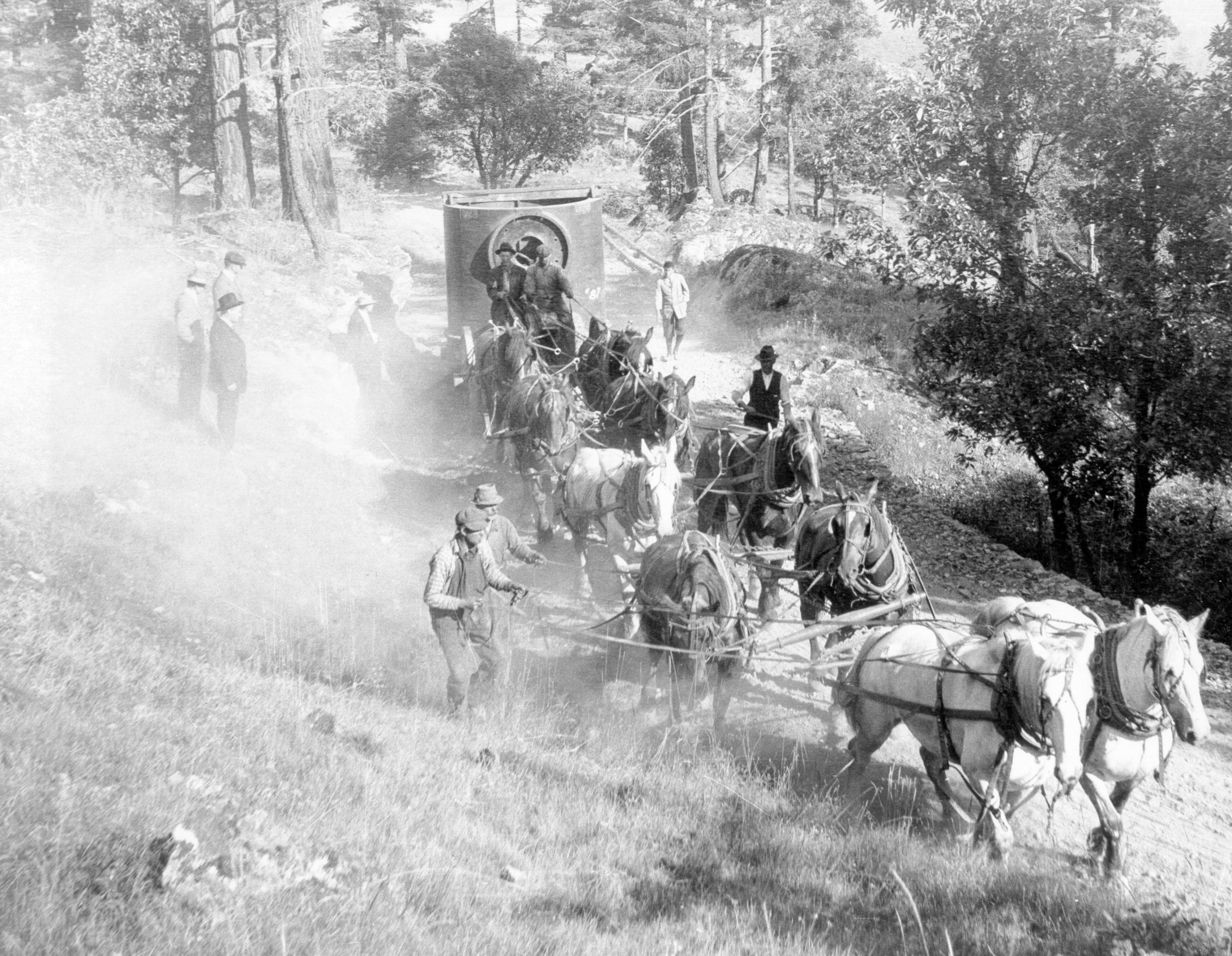 Photographie en noir et blanc d’un chariot tiré par des chevaux transportant un très gros équipement sur une route poussiéreuse à flanc de colline.