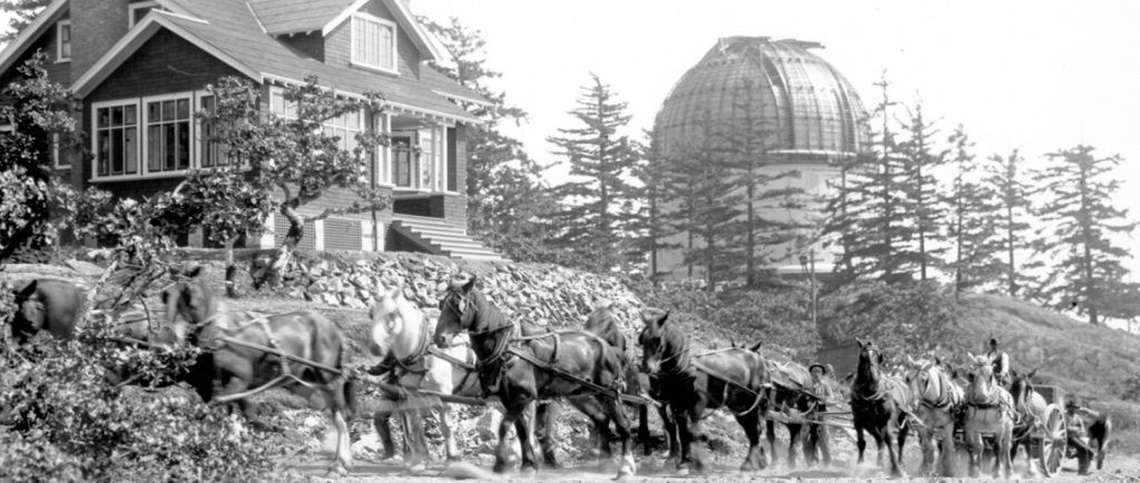 Photographie en noir et blanc montrant une partie du télescope transportée par un chariot tiré par des chevaux, avec la coupole du télescope et une maison à l’arrière-plan.
