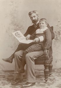 Photographie sépia d’un homme tenant un livre, assis sur une chaise, avec un jeune garçon assis sur ses genoux.