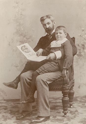 Photographie sépia d’un homme tenant un livre, assis sur une chaise, avec un jeune garçon assis sur ses genoux.