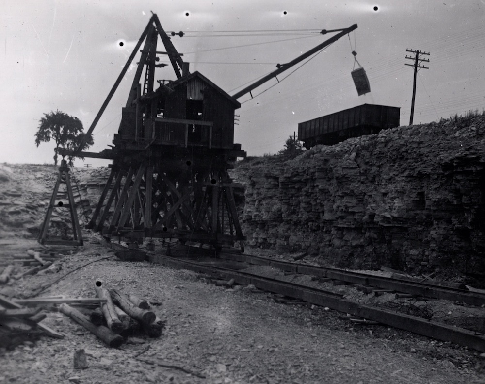 Image en noir et blanc d’une pièce d’équipement dans une carrière sur chenilles - L'appareil hisse une charge de fournitures sur un chariot ferroviaire en bordure de la carrière.