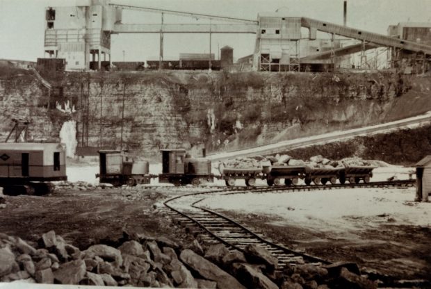 Un petit train et quatre charrettes sont vus le long d’une voie ferrée à l’intérieur d’une carrière. En arrière-plan se trouvent plusieurs structures industrielles.