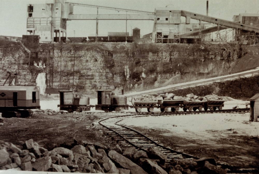 Un petit train et quatre charrettes sont vus le long d’une voie ferrée à l’intérieur d’une carrière. En arrière-plan se trouvent plusieurs structures industrielles.