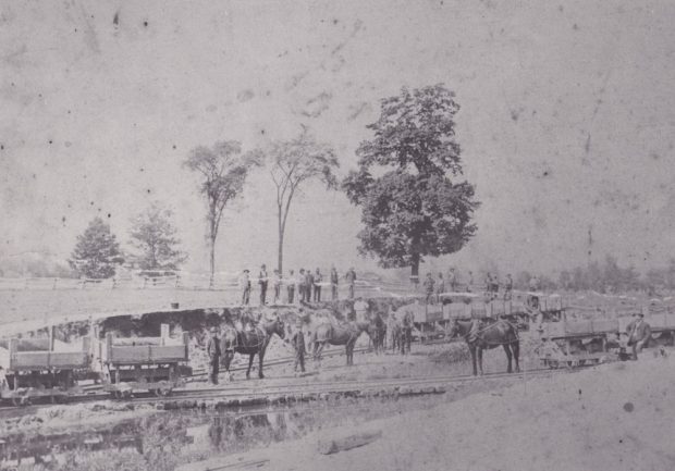  Une photographie en noir et blanc délavé d'une équipe de travail à l'extérieur, regardant un groupe de chevaux attachés à des charrettes en bois le long d'une piste