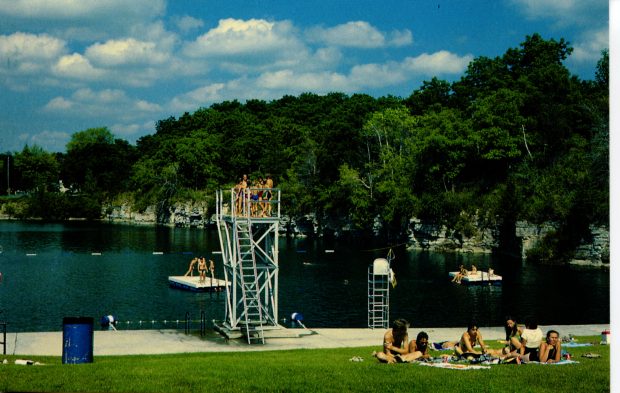 Des gens profitent d'une journée d'été au bord d'un lac de carrière : nager, attendre de sauter d'un plongeon en hauteur et se prélasser sur l'herbe. Il y a deux radeaux dans l’eau sur lesquels des personnes sont assises ou debout