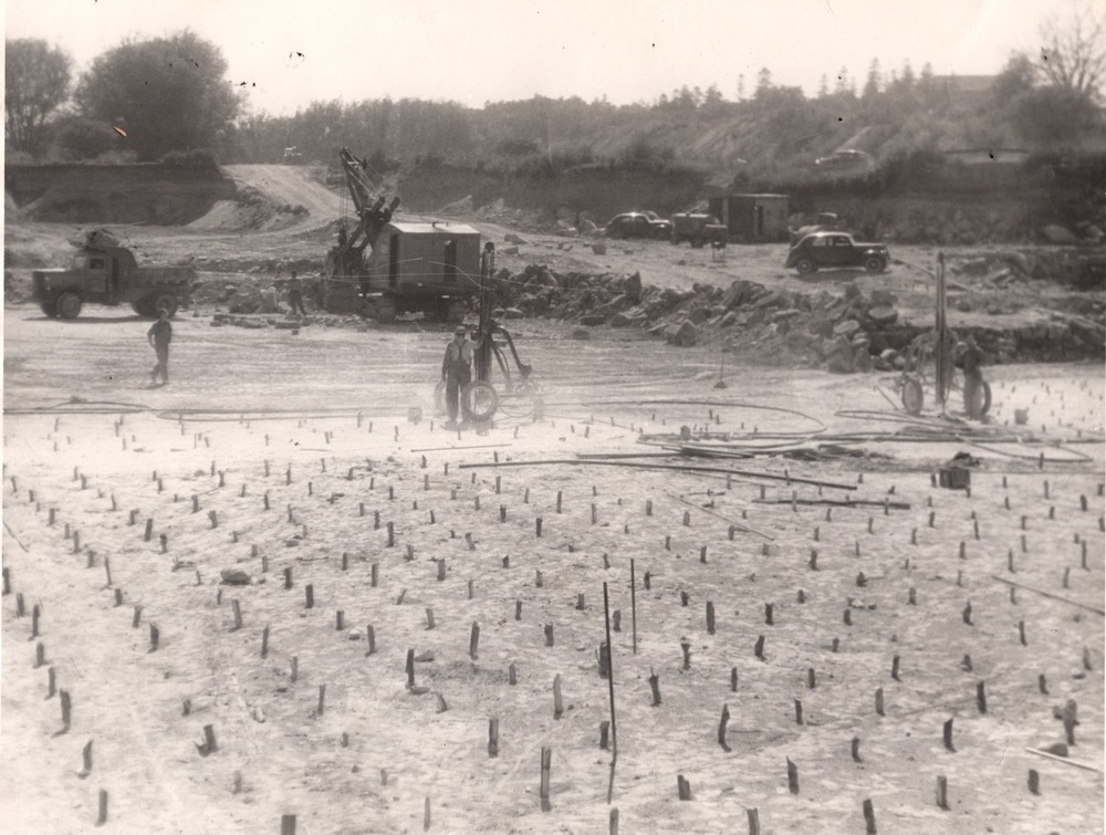 Des bâtons de dynamite sortent du sol en rangées. En arrière-plan se trouvent des voitures, des machines lourdes et des tas de pierres.