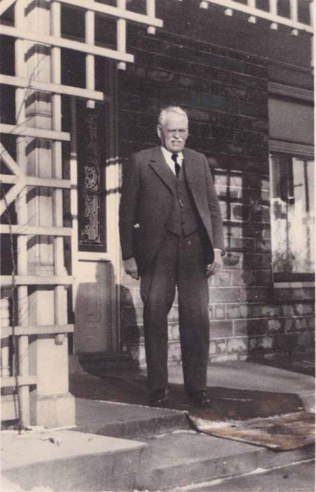 John Downing in a three-piece suit stands on a house's latticed porch squinting against the sun