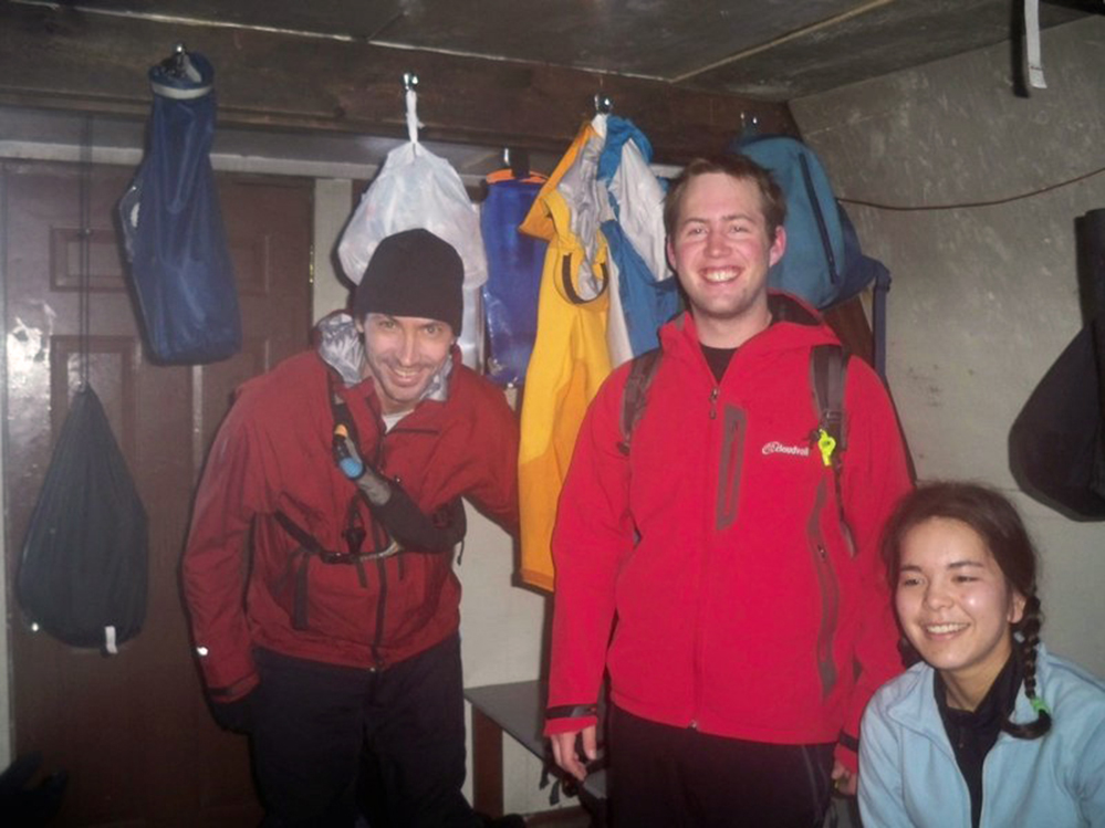 Trois membres du club posent pour la photo, deux hommes en vestes rouges et pantalons noirs sont debout tandis qu’une femme vêtue d’une fourrure bleu ciel est assise près de l’entrée principale.