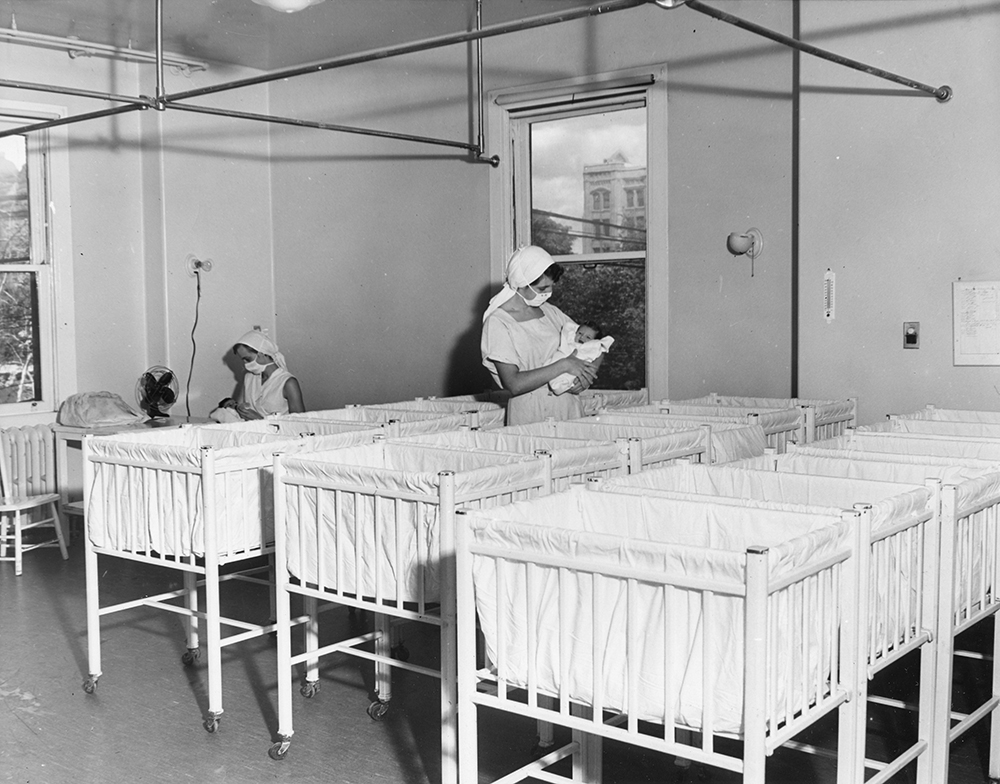 Deux infirmières portant des masques dans une pouponnière pleine de lits de bébé blancs dans une photo en noir et blanc. Chacune tient un nouveau-né.
