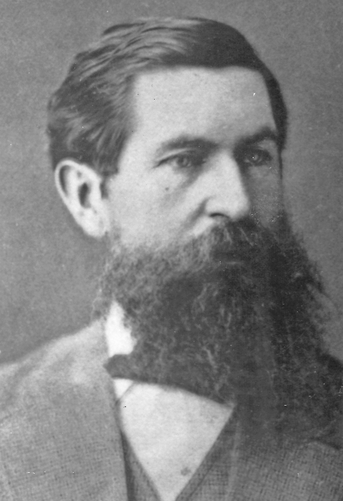 Portrait photographique en noir et blanc du docteur William Mostyn, un homme aux cheveux foncés avec une barbe, portant un costume.