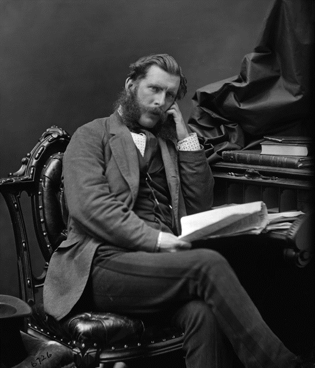 Portrait photographique en noir et blanc du Dr John Sweetland, âgé d'environ 35 ans, assis sur une chaise en cuir devant un bureau. Il porte un costume et tient un livre ouvert.