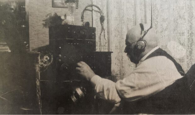 Photographie en noir et blanc d’un homme portant des écouteurs, assis sur une chaise à côté d’une radio. Il porte une chemise blanche et un gilet noir.