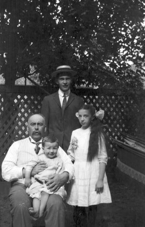 Photographie en noir et blanc du docteur Hanly, assis à l’extérieur avec ses enfants Arthur, debout derrière le docteur Hanly, Lois, debout à côté du docteur Hanly, et Bruce, assis sur les genoux du docteur Hanly.