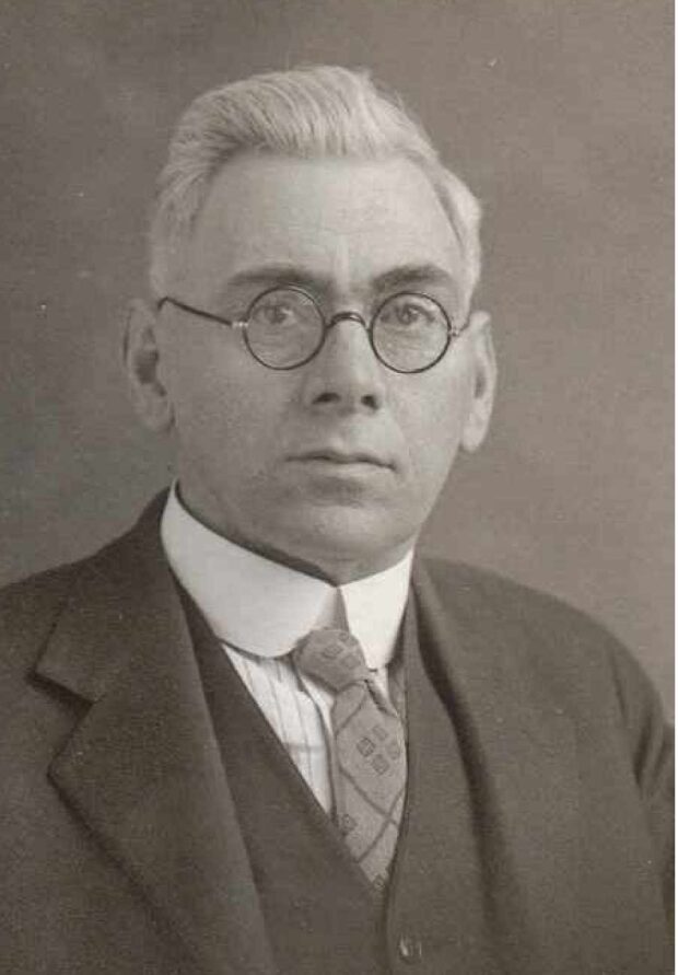 Portrait photographique en noir et blanc d'un homme aux cheveux gris qui porte des lunettes, une chemise blanche, une veste et une cravate.