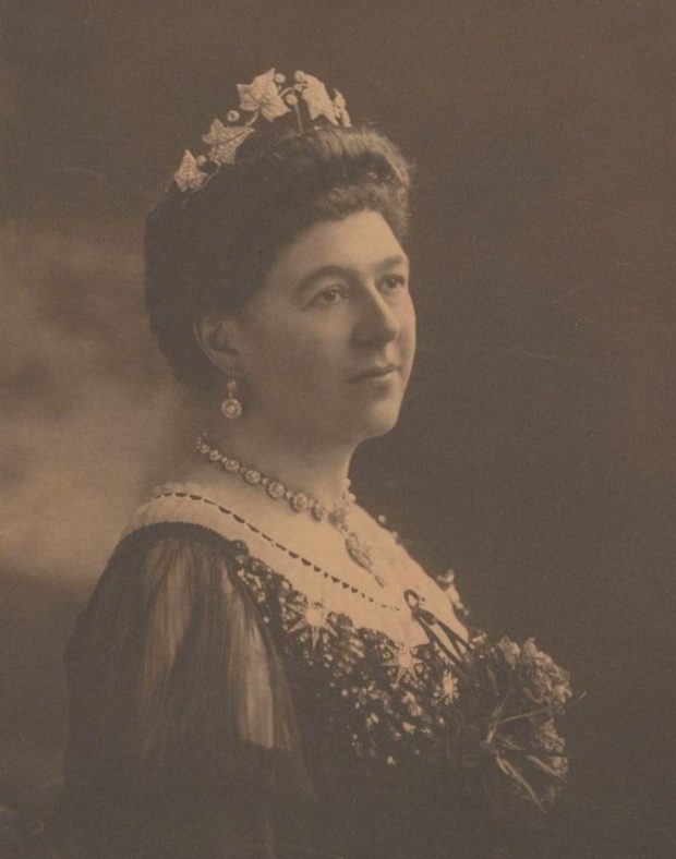 Photographie en sépia d'une femme aux cheveux foncés, portant une robe, un collier et des boucles d'oreilles. 