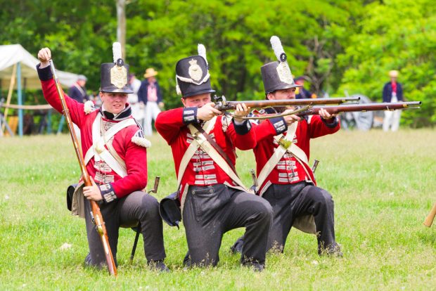 Photographie montrant trois comédiens en uniforme de soldat de 1812 à genoux. Deux sont en train de tirer, et l’autre recharge son fusil. 