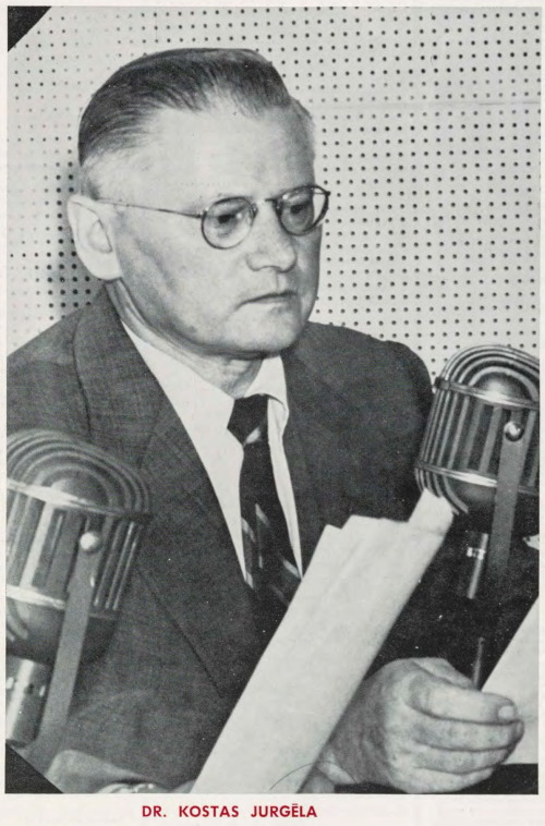 Photographie noir et blanc des années 1930 montrant un homme à lunettes qui parle dans un grand microphone à partir d’un texte, à l’intérieur d’une cabine d’enregistrement à panneaux perforés. 