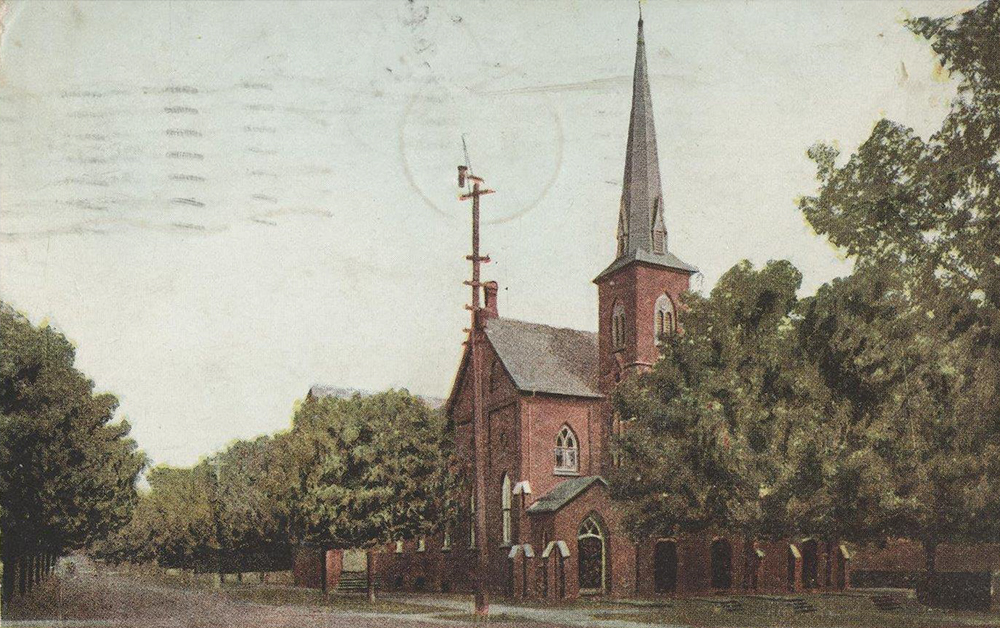 Carte postale colorisée d’une église orillienne sur une rue bordée d’arbres, vers 1909. L’église a un seul clocher et est faite en brique rouge.