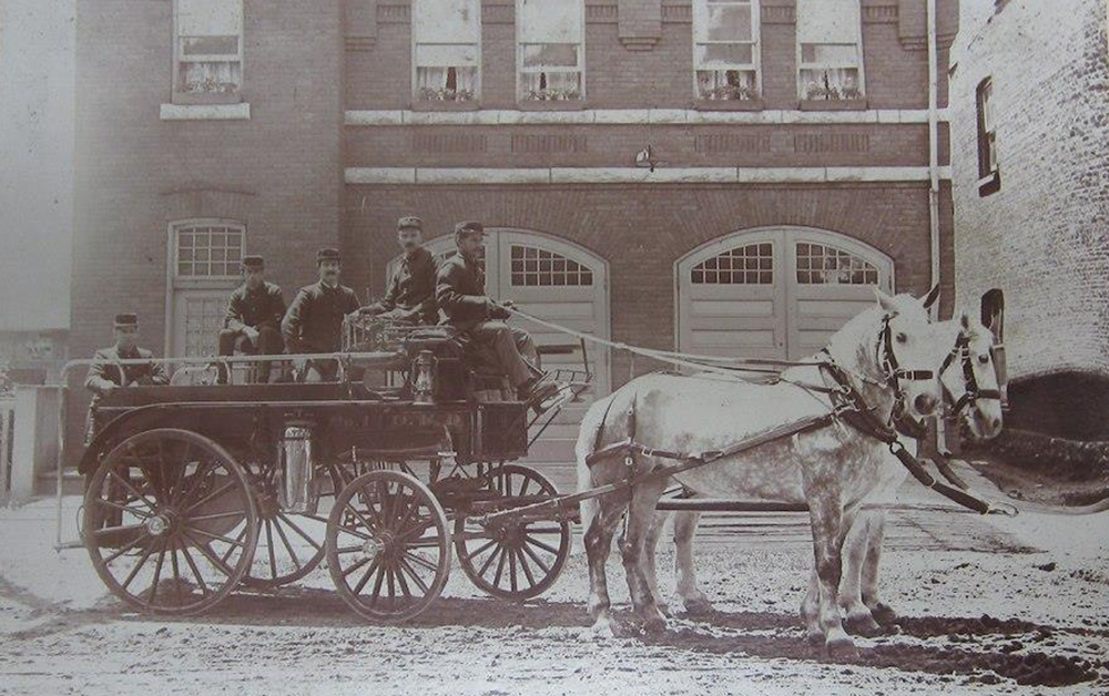 Photo sépia prise en 1900 du corps de pompiers d’Orillia devant la caserne en brique. Cinq hommes sont assis sur un chariot d’incendie auquel sont attelés deux chevaux blancs sur une route de terre.