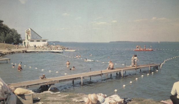 Carte postale d’environ 1960 montrant la plage Couchiching parsemée de gens assis sur les quais, en train de nager ou de se faire bronzer. À l’arrière-plan se trouve l’iconique Aqua Theatre d’Orillia.