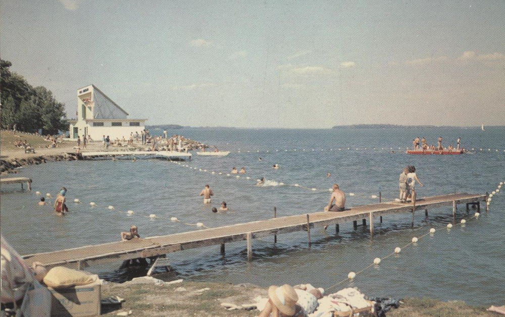 Carte postale d’environ 1960 montrant la plage Couchiching parsemée de gens assis sur les quais, en train de nager ou de se faire bronzer. À l’arrière-plan se trouve l’iconique Aqua Theatre d’Orillia.