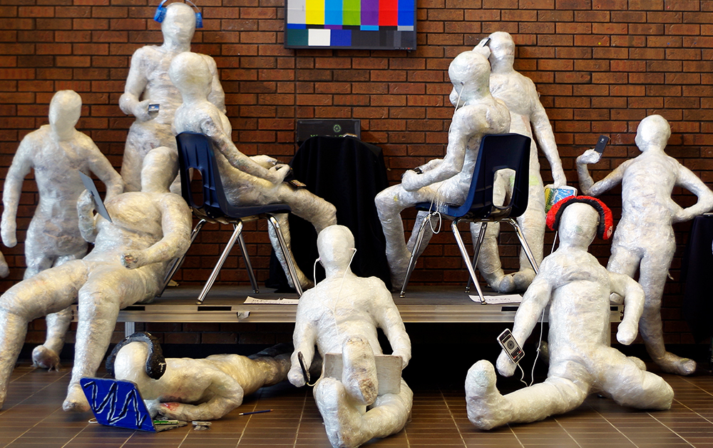 Dix sculptures blanches à échelle humaine représentent des gens disposés devant un mur de brique rouge. Chaque sculpture interagit avec un appareil électronique et non avec les autres.