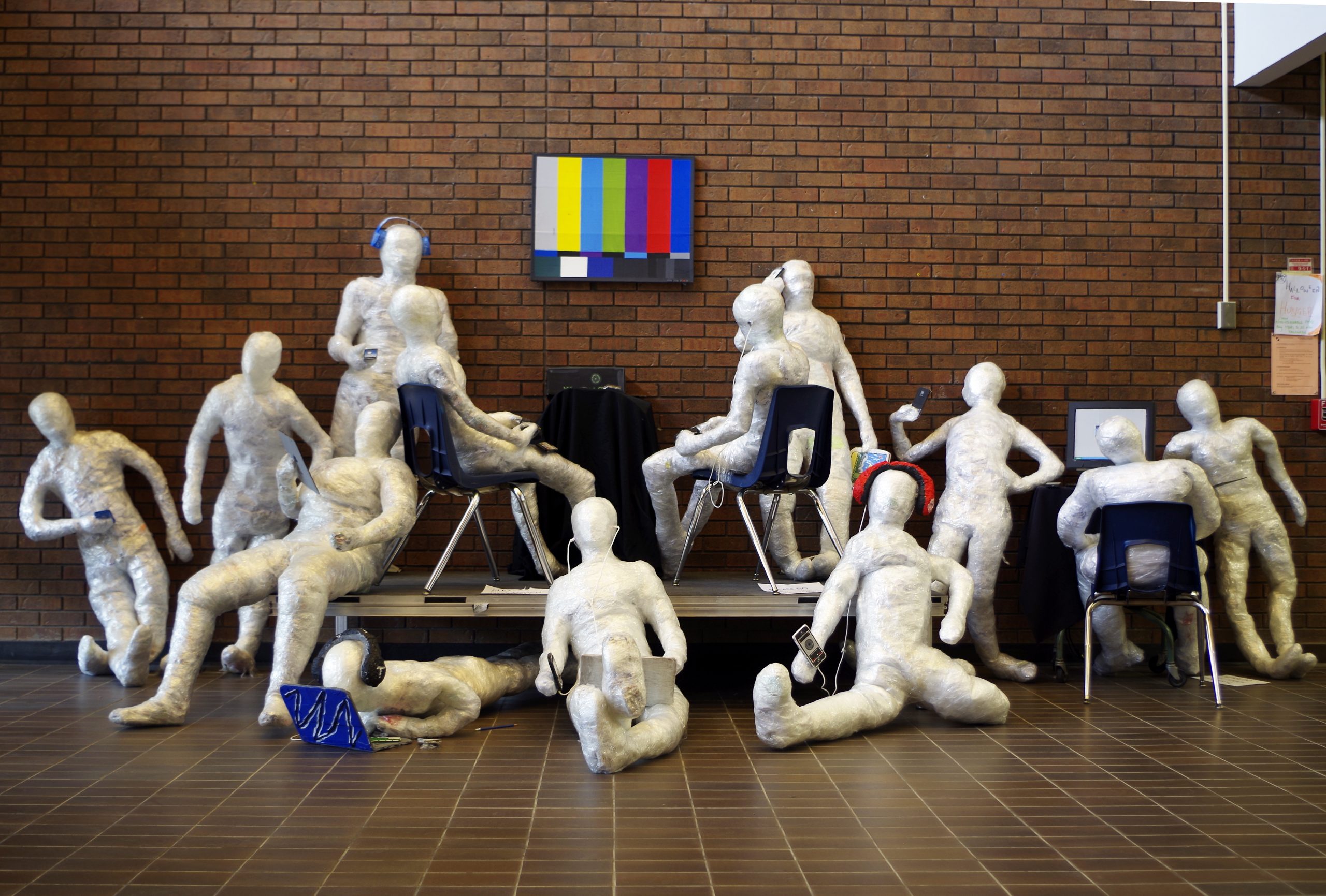 Dix sculptures blanches à échelle humaine représentent des gens disposés devant un mur de brique rouge. Chaque sculpture interagit avec un appareil électronique et non avec les autres.