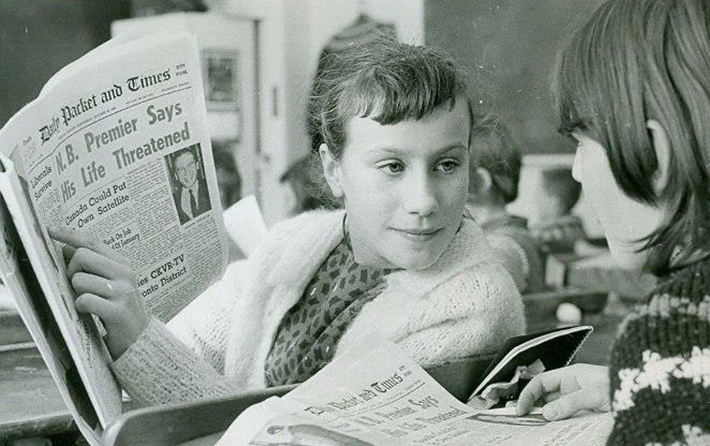 Photo en noir et blanc d’environ 1960 représentant deux écolières lisant le Packet & Times en classe. L’une d’elles indique du doigt un article, que l’autre regarde.