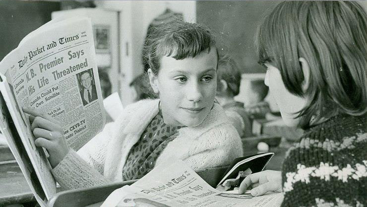 Photo en noir et blanc d’environ 1960 représentant deux écolières lisant le Packet & Times en classe. L’une d’elles indique du doigt un article, que l’autre regarde.