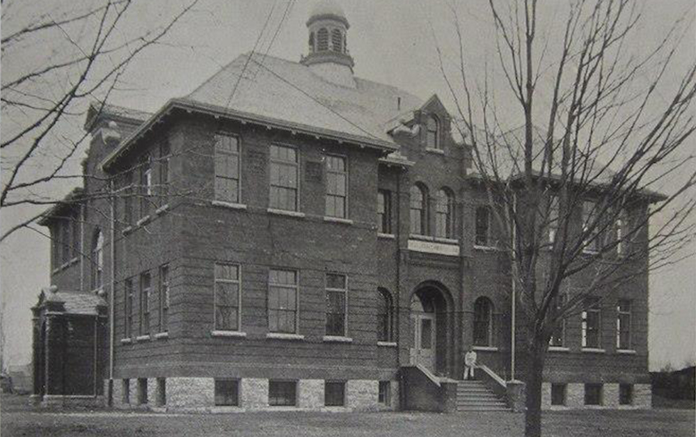 Photo en noir et blanc d’un homme habillé en blanc assis à l’entrée d’un grand bâtiment en brique. Sous la photo se trouve l’inscription en anglais : Collège d’Orillia, Rue Borland, 1899.