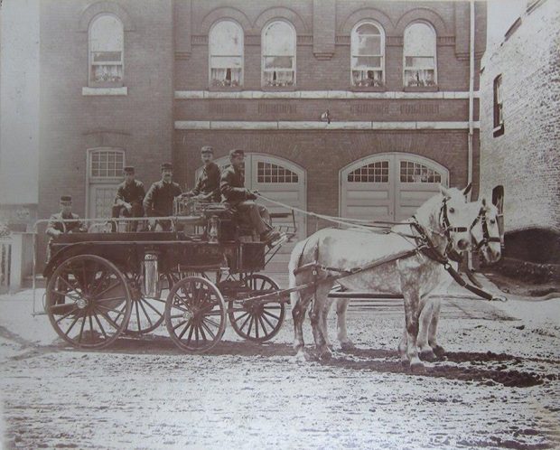 Photo sépia prise en 1900 du corps de pompiers d’Orillia devant la caserne en brique. Cinq hommes sont assis sur un chariot d’incendie auquel sont attelés deux chevaux blancs sur une route de terre.