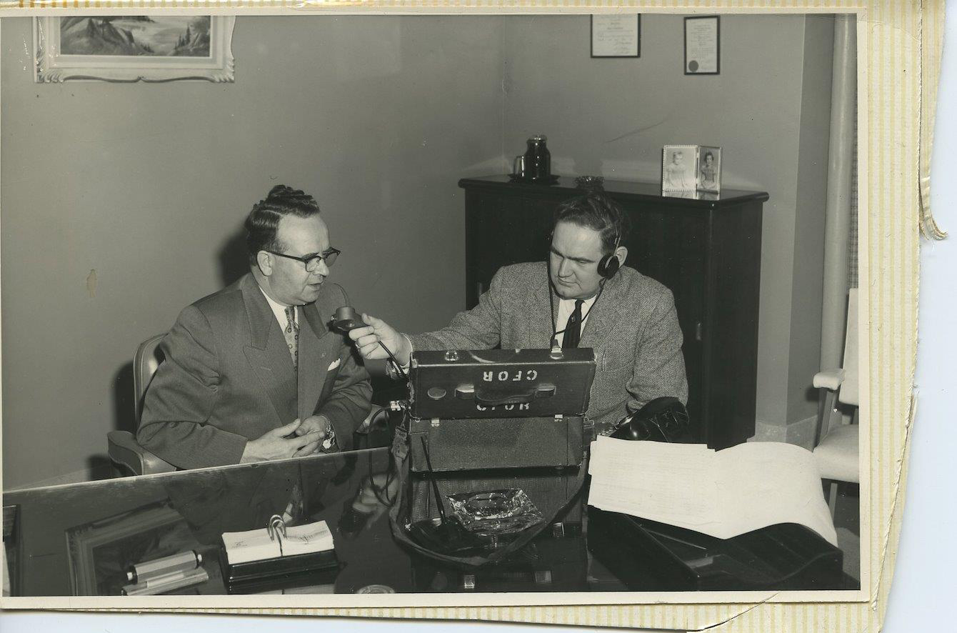 Photo en noir et blanc aux bords jaunis et déchirés datant d’environ 1950. Pete McGarvey, assis à un grand bureau avec du matériel d’enregistrement de CFOR, tend un micro vers un homme bien habillé avec des lunettes à monture noire.