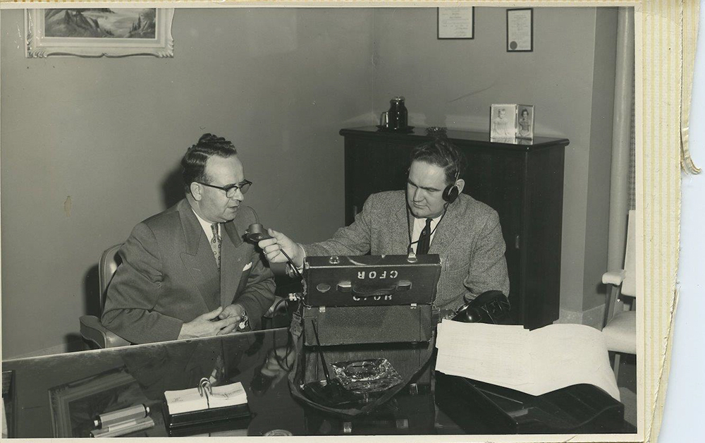 Photo en noir et blanc aux bords jaunis et déchirés datant d’environ 1950. Pete McGarvey, assis à un grand bureau avec du matériel d’enregistrement de CFOR, tend un micro vers un homme bien habillé avec des lunettes à monture noire.