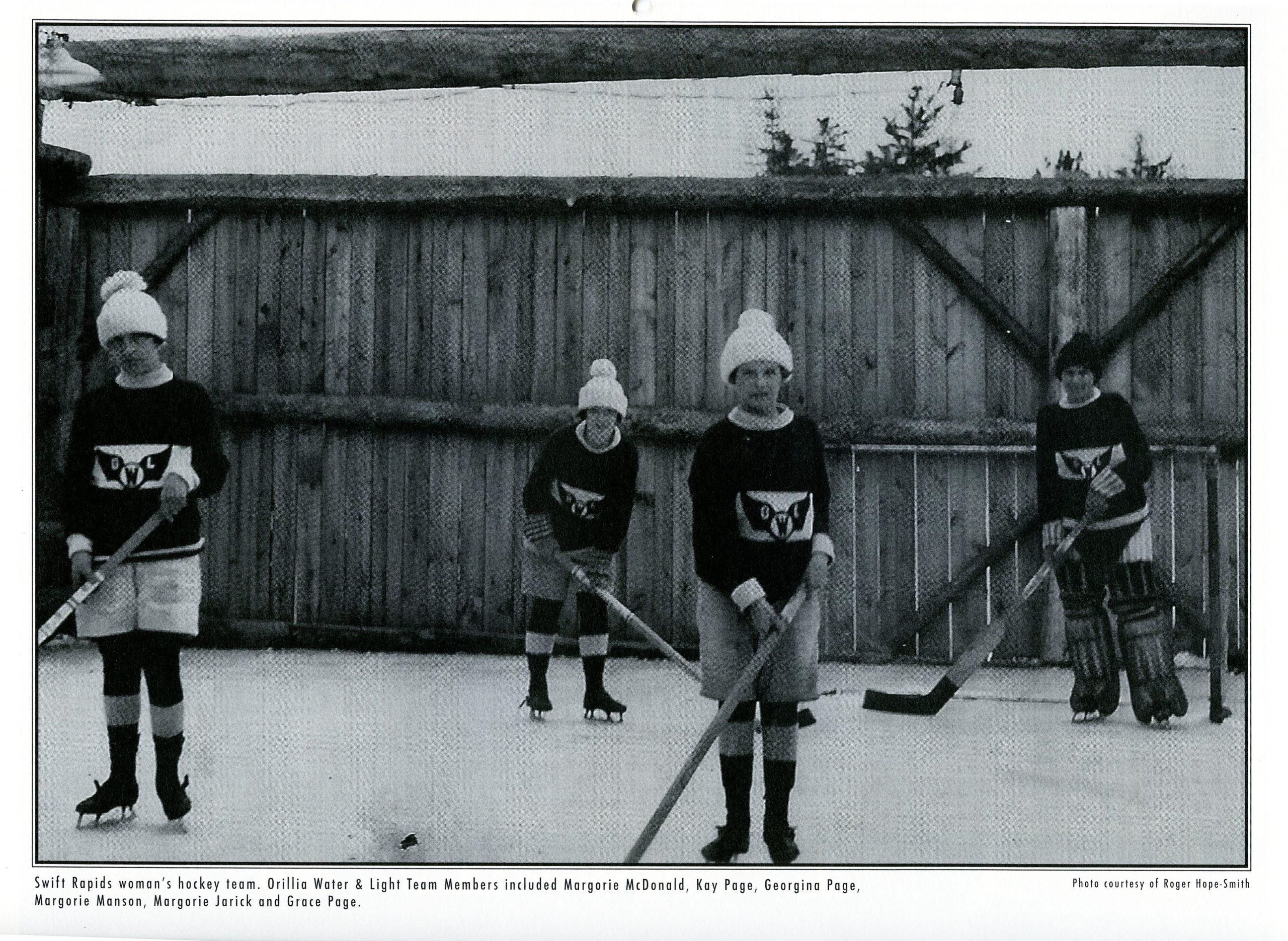 Photo en noir et blanc datant d’environ 1925. Quatre jeunes filles sont chaudement habillées dans leur uniforme de hockey, bâton à la main, sur une patinoire extérieure devant une haute clôture en bois.