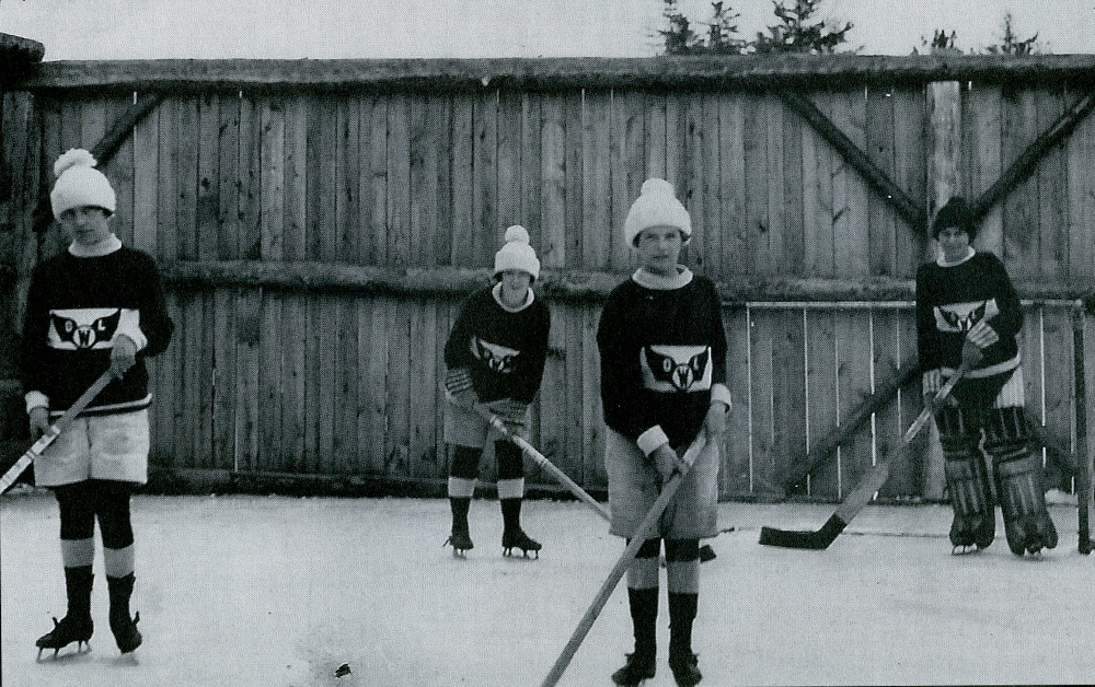 Photo en noir et blanc datant d’environ 1925. Quatre jeunes filles sont chaudement habillées dans leur uniforme de hockey, bâton à la main, sur une patinoire extérieure devant une haute clôture en bois.