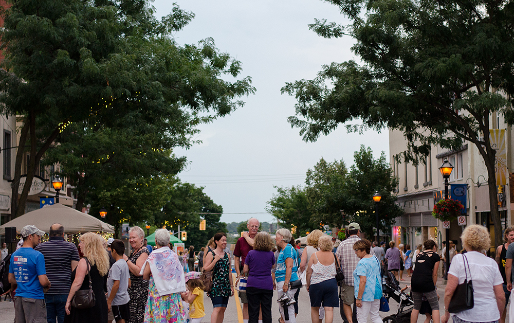 Lors d’une chaude soirée d’août en 2016, des gens discutent sur une rue achalandée du centre-ville d’Orillia, bordée d’arbres à feuilles vertes, de lampadaires allumés et de bâtiments historiques.