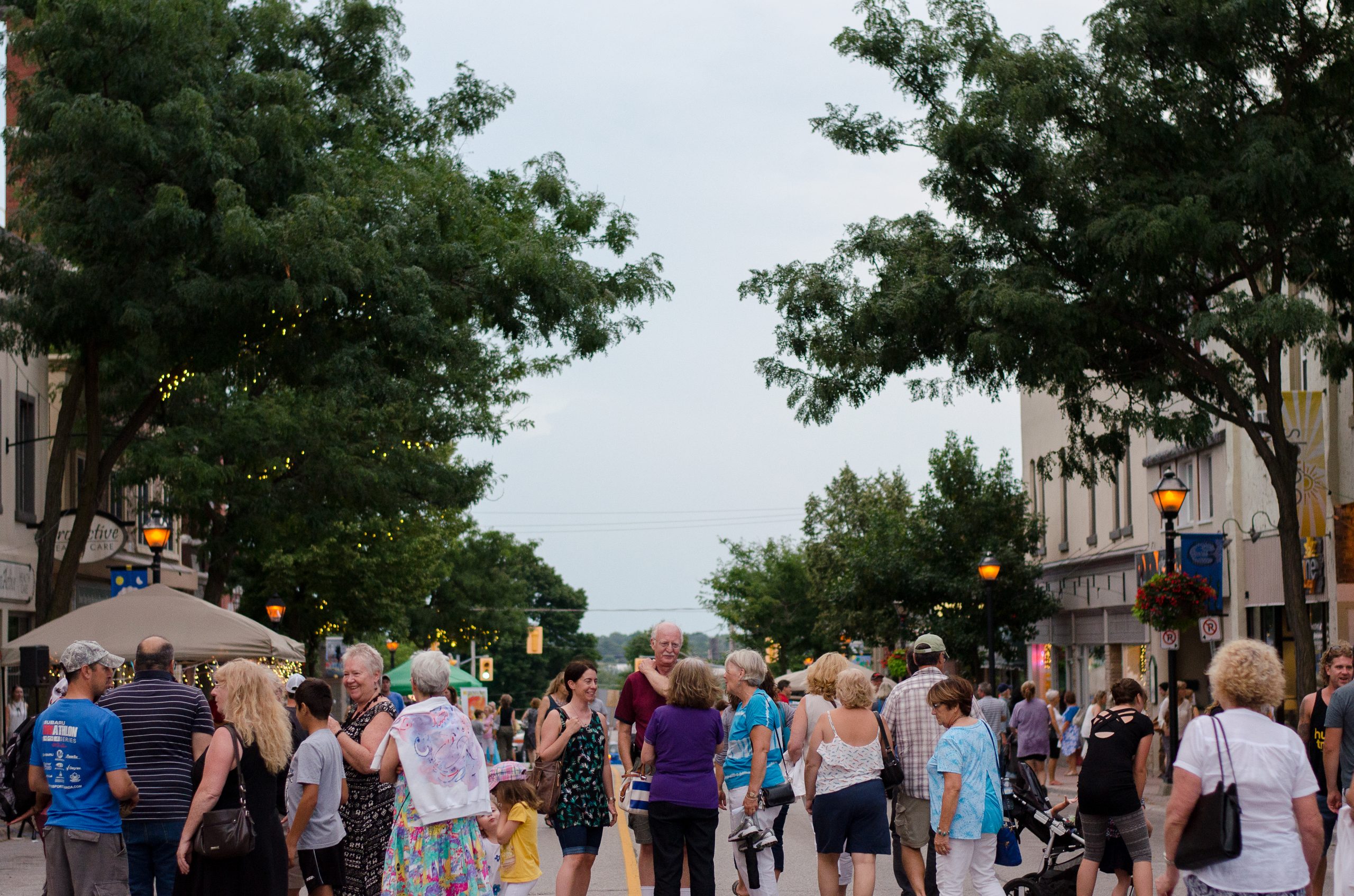 Lors d’une chaude soirée d’août en 2016, des gens discutent sur une rue achalandée du centre-ville d’Orillia, bordée d’arbres à feuilles vertes, de lampadaires allumés et de bâtiments historiques.