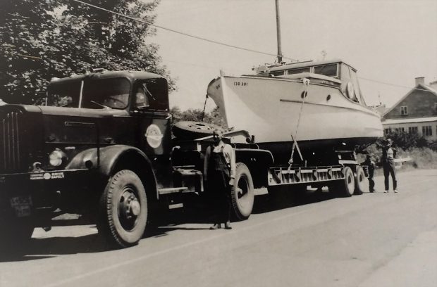 Photographie noir et blanc d’un yacht blanc, posé sur une remorque, attachée à un camion noir. F-X Lachance est debout à la gauche du camion. « Le Notre-Dame, 1952. appartenant au Curé Bélanger de Berthier »