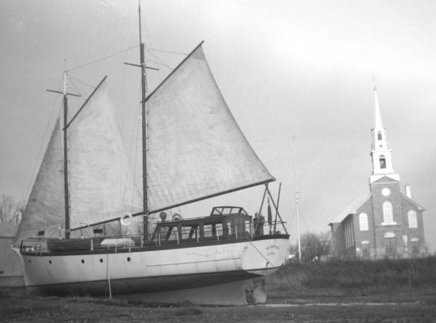 Photographie noir et blanc d’un voilier à deux mats, voiles déployées, sur la grève près de l’église de Saint-Laurent. « L’Intrépide, 1932. Robert Taschereau »