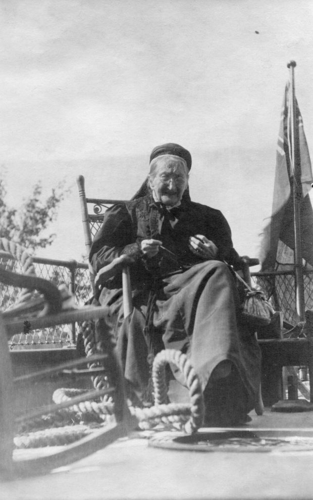 Une vieille femme assise sur une chaise et qui tricote sur le pont d’un bateau-logement, portant une robe noire.