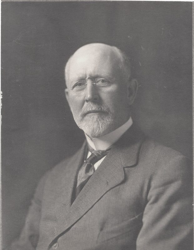 Photographie de studio d’un homme aux cheveux blancs qui porte un complet noir.