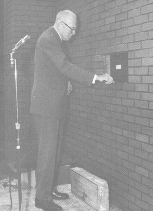 Un homme portant des lunettes, debout derrière un microphone, sur le point de poser une brique dans le mur d’un bâtiment.