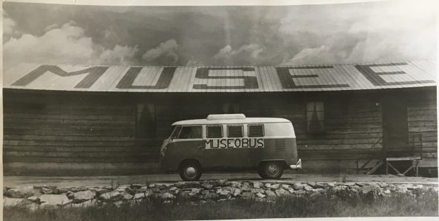 Photo en noir et blanc d'une fourgonnette identifée Muséobus stationnée devant un bâtiment sur lequel est inscrit, en grosses lettres, le mot Musée. 