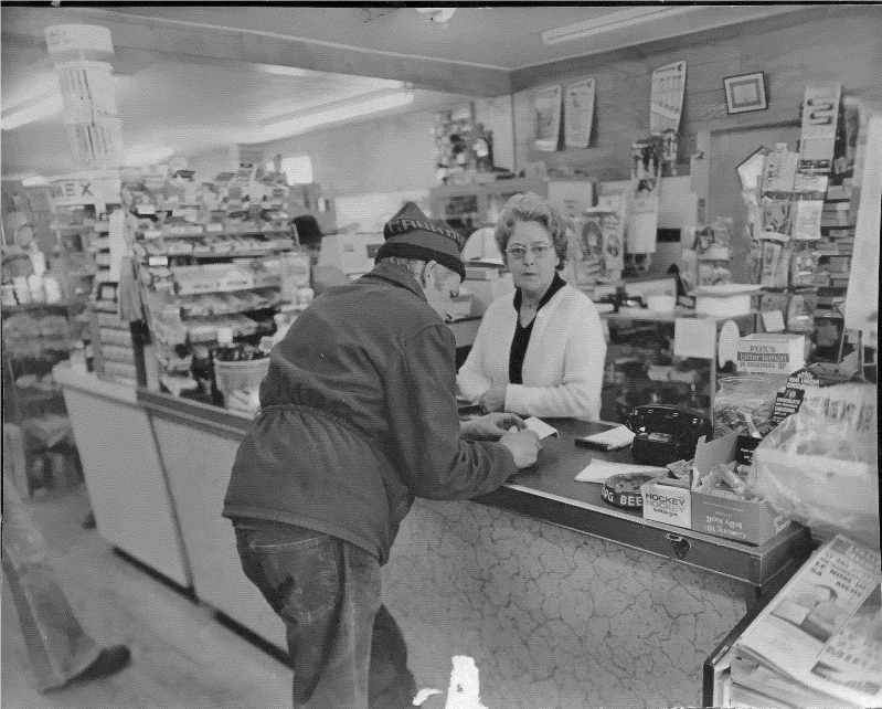 Photo en noir et blan d'une femme et d'un homme au comptoir caisse d'un magasin général. 