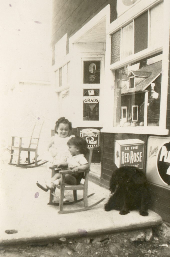 Photo en noir et blanc de deux enfants et un chien devant une épicerie.