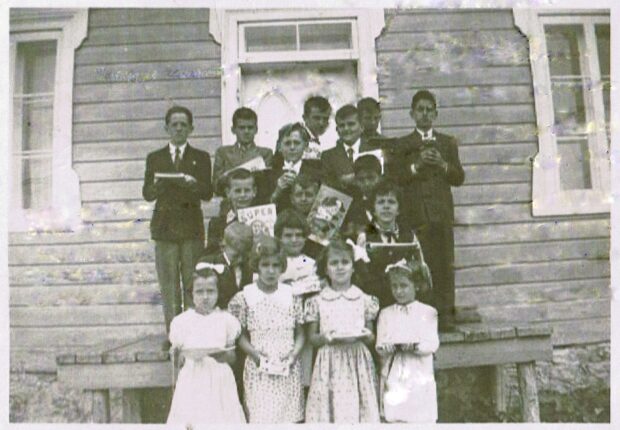 Photo en noir et blanc d'un groupe d'enfants tenant un livre devant l'école. 